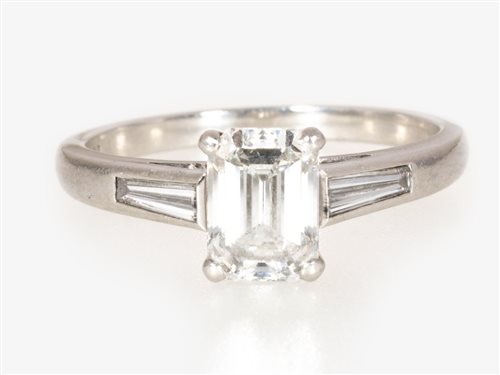 Lot 174 - A diamond solitaire ring, the emerald cut stone four claw set in a platinum mount with a tapered baguette cut diamond set into each shoulder