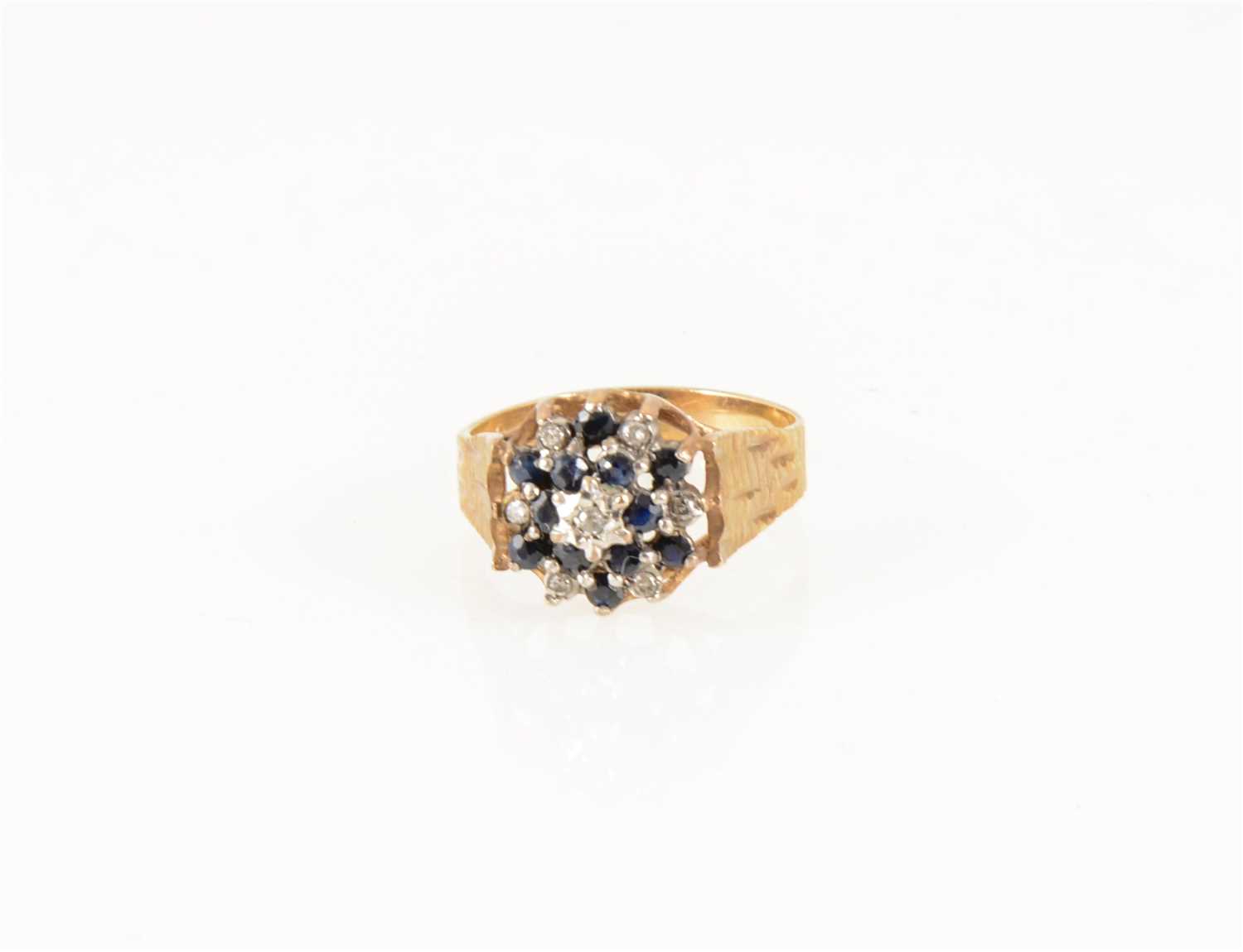 Lot 176 - A sapphire and diamond circular cluster ring, twelve sapphires claw set, seven diamond points illusion set in a 9 carat yellow and white gold mount with wide bark textured shoulders